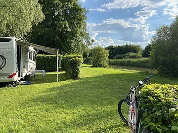 AZUR Campingplatz Kipfenberg Auf dem Campinplatz steht links ein Wohnmobil mit herausgelassener Jalousie. Hinter dem Mobil ragen große Bäume auf. Die Wiesen und Sträucher strahlen im hellen grün. Rechts lehnt ein Fahrrad an einem Strauch.