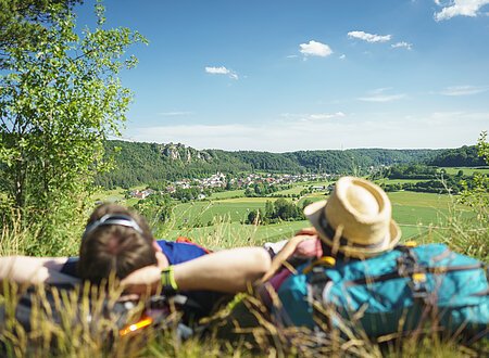 Ein Mann mit verschränkten Armen und daneben eine Frau mit Strohhut, die ihren Kopf auf ihren blauen Rucksack abgelegt hat, liegen im Gras. Sie genießen den Blick von der Arnsberger Leite ins Tal. Der Himmel strahlt blau und nur ein paar Wolken haben sich dazu verirrt.