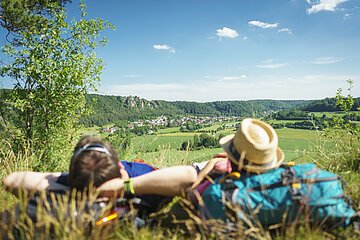 Ein Mann mit verschränkten Armen und daneben eine Frau mit Strohhut, die ihren Kopf auf ihren blauen Rucksack abgelegt hat, liegen im Gras. Sie genießen den Blick von der Arnsberger Leite ins Tal. Der Himmel strahlt blau und nur ein paar Wolken haben sich dazu verirrt.