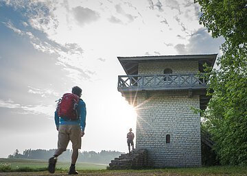 Ein Wanderer mit rotem Rucksack und blauer Jacke geht zum Limesturm. Auf den Treppen des Limesturm steht eine Frau und wartet. Der Limesturm ist komplett restauriert und ist aus weißem Stein gebaut. Oberhalb ist ein Geländer mit kleinem Rundgang aus Holz angebracht. Der Himmel ist bewölkt.