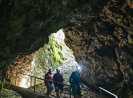 Drei Wanderer steigen die Treppenstufen zur Höhle hinab. Der Vorderste ist mit einer Stirnlampe ausgestattet und leuchtet ihnen den Weg aus. Licht kommt nur vom Eingang in die Höhle.