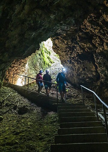 Drei Wanderer steigen die Treppenstufen zur Höhle hinab. Der Vorderste ist mit einer Stirnlampe ausgestattet und leuchtet ihnen den Weg aus. Licht kommt nur vom Eingang in die Höhle.