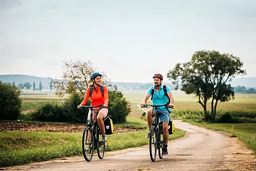 Eine Frau und ein Mann radeln auf dem Altmühltal-Radweg bei Dittenheim. Beide tragen einen Fahrradhelm und einen Rucksack. Der Radweg wird von Gras und grünen Bäumen gesäumt.