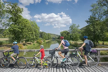 Eine Familie mit dem Rad unterwegs hält an einer Brücke am Ludwig-Donau-Main-Kanal bei Altessing. Das Ufer ist von grünen Bäumen gesäumt und der Himmel strahlt hellblau.