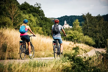 Zwei Radler, eine Frau und ein Mann, sind auf dem Radweg unterwegs. Die Gräser entlang des Radweges sind recht schon recht vertrocknet und braun. Ihr Ziel ist die Burg Kipfenberg die inmitten des Waldes in weiß bereits unscharf zu sehen ist.