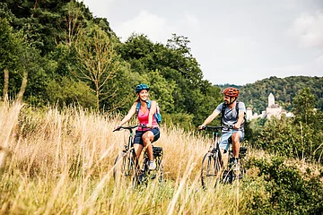 Zwei Radler, eine Frau und ein Mann, sind auf dem Radweg unterwegs. Die Gräser entlang des Radweges sind recht schon recht vertrocknet und braun. Dahinter blitzt in weiß die Burg Kipfenberg inmitten des Waldes heraus.