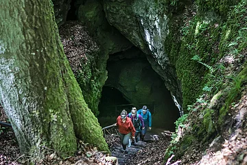 Aus dem Eingang der Höhle sind drei Wanderer nach oben unterwegs. Die Höhle liegt mitten im Wald und der felsige Eingang ist mit Moos übersät.