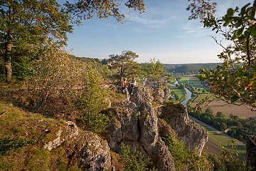 Ein Wanderpärchen, etwas entfernt, steht links auf einem Felsen an der Arnsberger Leite, die gut zu sehen ist. Dahinter erstreckt sich der Blick ins Tal. Der Himmel erstrahlt wolkenlos.