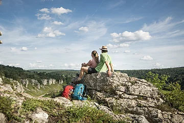 Ein Pärchen sitzt auf einem Felsen. Ihre Wanderrucksäcke lehnen daran. Sie lassen ihren Blick ins Tal schweifen.