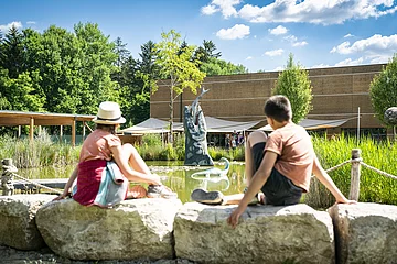 Zwei Kinder sitzen am Wasser im Außengelände des Dinosauriermuseum und beobachten die grüne Landschaft und das Wasser.