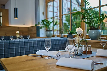 Ein gedeckter Holztisch mit weißen Servietten ist im Vordergrund. Die Bank ist mit blau-kariertem Stoff bezogen, rechts kann man zum Fenster hinausschauen.