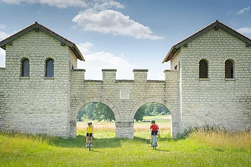 Zwei Gravelbiker einer mit gelben und einer mit roten Shirt fahren durch die Tore des rekonstruierten Kastells auf der Wiese.