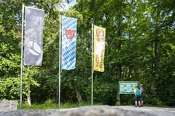 Zwei Wanderer stehen an der Infotafel am Geographischen Mittelpunkt Bayerns bei Kipfenberg und lesen interessiert. Links von ihnen sind drei Fahnen gehisst.