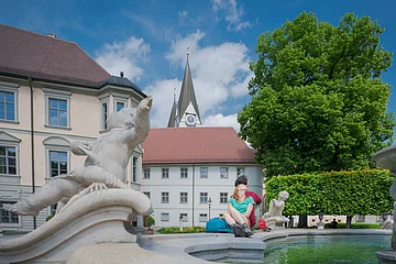 Ein Pärchen sitzt am Rand des Brunnens und genießt den Flair des Platzes. Links ist das Landratsamt teilweise zu erkennen. Die Kirchentürme ragen über ein Gebäude heraus.