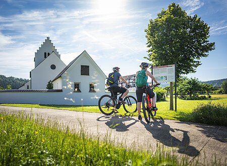 Radfahrer vor einer Infotafel zum obergermanisch-raetischen Limes an der Kirche St. Johannes in Böhming. Im Hintergrund ist das Mesnerhaus (Fachwerkhaus) zu sehen.