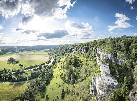 Weiter Blick ins Altmühltal: Rechts Felsen mit grünem Hang ins Tal  und rechts sieht man den Fluss im Tal mit Wiesen.
