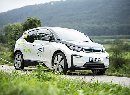 e-altmuehltal | mein Elektroauto Ein weißes Elektroauto mit SGD Pharma-Logo fährt auf einer ländlichen Straße, im Hintergrund sind Bäume und Hügel.