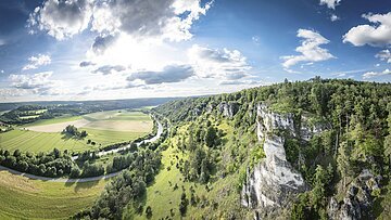 Landschaft mit grünen Feldern, einem Fluss und bewaldeten Hügeln unter einem bewölkten Himmel.
