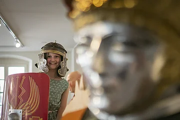 Kind trägt römischen Helm und hält Schild, im Hintergrund eine Statue.