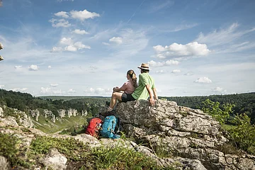 Zwei Wanderer sitzen auf einem Felsen und blicken auf bewaldete Hügel unter blauem Himmel mit Wolken.