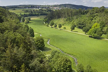 Tal mit grünem Gras, Bäumen, kleinem Bach und Dorf im Hintergrund unter blauem Himmel