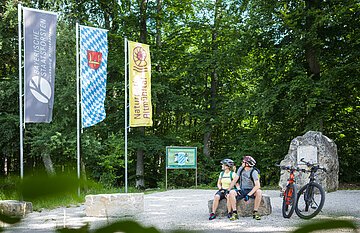 Zwei Radfahrer mit Helm sitzen auf Steinbank im Wald neben zwei Mountainbikes und drei Fahnen.