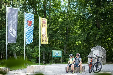 Zwei Radfahrer mit Helm sitzen auf Steinbank im Wald neben zwei Mountainbikes und drei Fahnen.