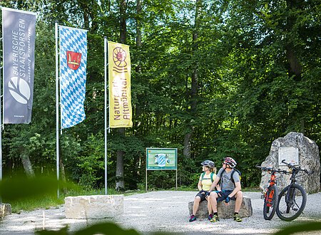 Geographischer Mittelpunkt Bayerns Zwei Radfahrer mit Helm sitzen auf Steinbank im Wald neben zwei Mountainbikes und drei Fahnen.