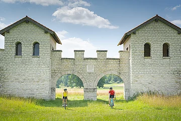 Zwei Radfahrer fahren durch zwei Bögen einer historischen Steinmauer in einer grünen Wiesenlandschaft.