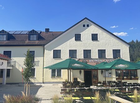 Außenansicht eines gelben Gasthofs mit grünen Sonnenschirmen und bestuhltem Biergarten bei Sonnenschein.