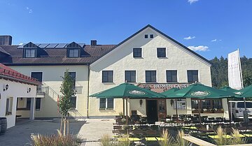 Außenansicht eines gelben Gasthofs mit grünen Sonnenschirmen und bestuhltem Biergarten bei Sonnenschein.