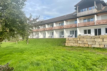 Mehrstöckiges Wohngebäude mit Balkonen, davor eine grüne Wiese und ein Baum links im Bild.