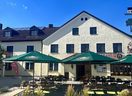Außenansicht eines Gasthauses mit grünen Sonnenschirmen und Sitzgelegenheiten bei klarem Himmel.