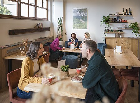 Ein junges Paar sitzt sich am Tisch im Schokoladencafé Rose Kakao in Beilngries gegenüber und isst Kuchen sowie trinkt heiße Schokolade und Tee. In einer Sitzecke genießt eine Gruppe von drei Freundinnen ebenfalls Kuchen und heiße Getränke.