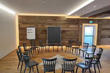 Konferenzraum mit Holzfußboden, Stühlen im Kreis, Flipchart mit „herzlich willkommen“ und Holzwand mit Tür und Tafel.