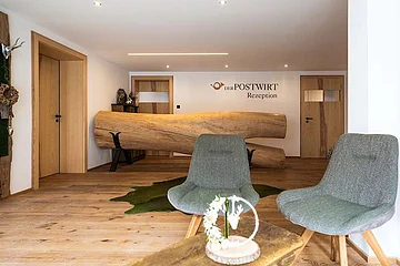 Empfangsbereich mit rustikalem Holztresen, zwei grauen Stühlen und Holzboden, Wand mit Schriftzug „Der Postwirt Rezeption“