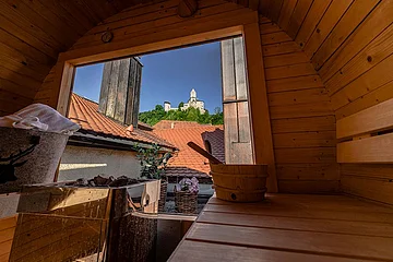 Blick aus einer Holzsauna auf ein geöffnetes Fenster mit Dachziegeln und einer Burg auf einem Hügel im Hintergrund.