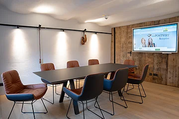 Konferenzraum mit sechs braunen Stühlen um schwarzen Tisch, Holzwand und Bildschirm an weißer Wand.