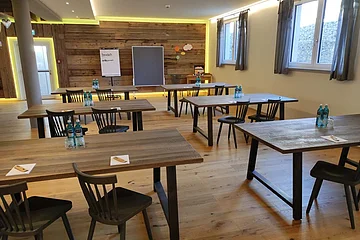 Konferenzraum mit Holztischen, Stühlen, Wasserflaschen und Flipchart vor Holzpaneelwand und Fenstern mit Vorhängen.