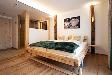 Schlafzimmer mit Holzbett, grünem Überwurf, Holzboden und offener Dusche hinter Holztrennwand.