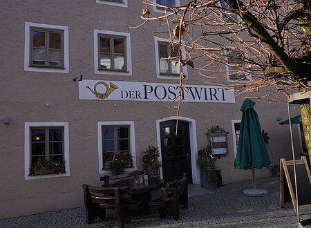 Außenansicht eines Gebäudes mit Schild „Der Postwirt“, Tischen, Stühlen und kahlem Baum im Vordergrund.