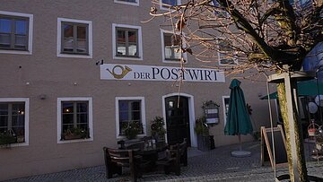 Außenansicht eines Gebäudes mit Schild „Der Postwirt“, Tischen, Stühlen und kahlem Baum im Vordergrund.
