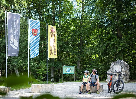 Geographischer Mittelpunkt Bayerns Zwei Radfahrer mit Helm sitzen auf Steinbank im Wald neben zwei Mountainbikes und drei Fahnen.