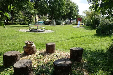 Spielplatz mit Baumstamm-Sitzkreis, Karussell, Klettergerüst und Rutsche auf einer Wiese mit Bäumen im Hintergrund